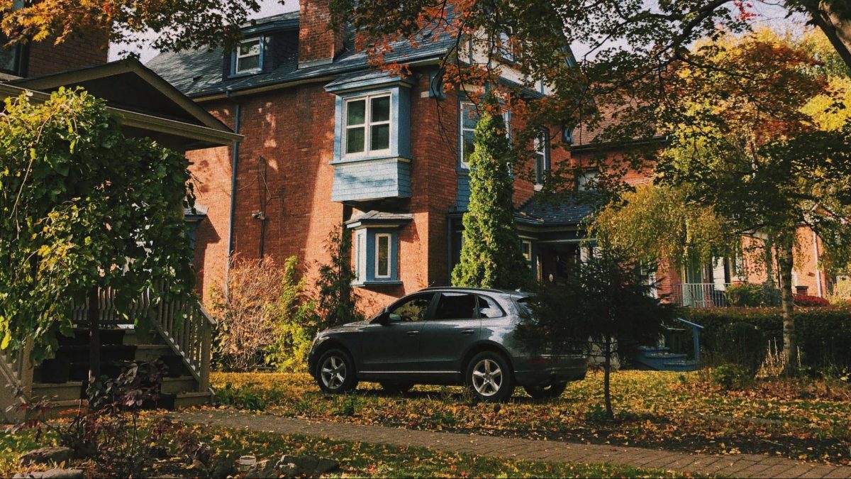 A car parked on the driveway of a classic brick house with autumn foliage.