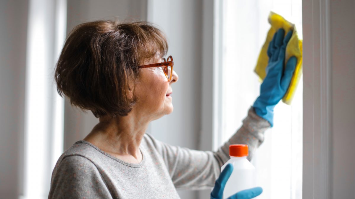 A senior woman with glasses is cleaning a window, wearing blue gloves and holding a yellow cloth and a spray bottle, illustrating diligent home maintenance.