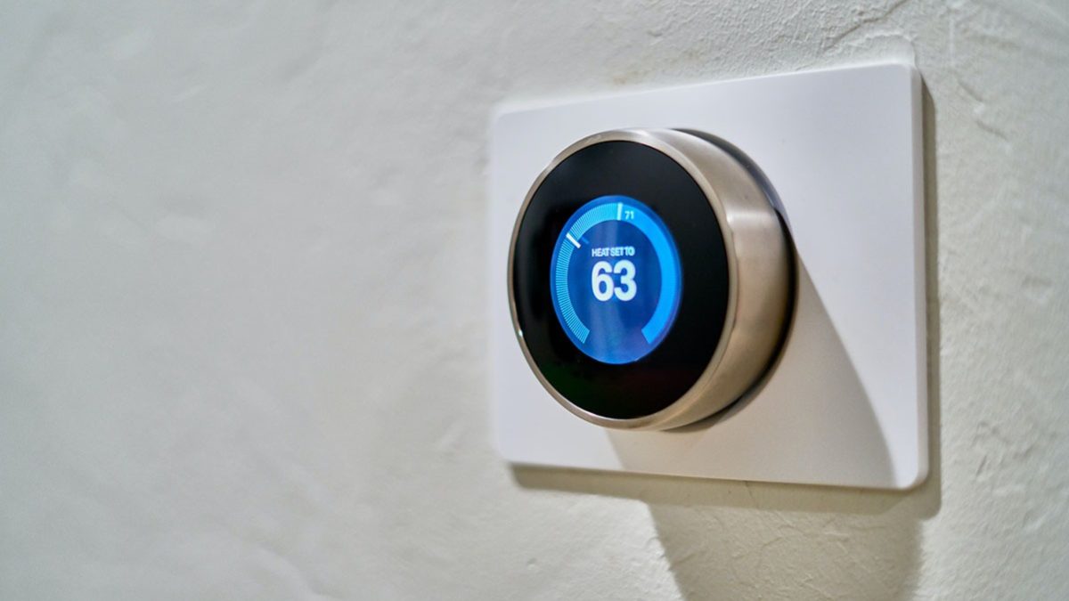 A close-up view of a round digital thermostat with a blue-lit display showing a temperature setting of 63 degrees, mounted on a textured white wall.