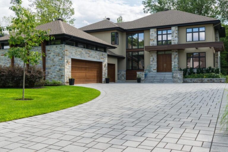 A modern, two-story home with a beautifully landscaped front yard. The house features a spacious driveway made of interlocking stone pavers, a double garage with wooden doors, and large windows that provide ample natural light. The exterior walls are a combination of stone and stucco, giving the home a stylish and contemporary look.